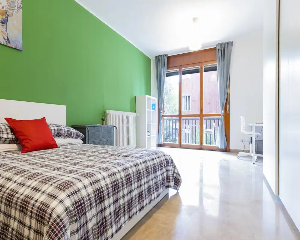 Bright, modern bedroom with a large bed in the foreground, a green accent wall and floor-to-ceiling window letting in abundant natural light.