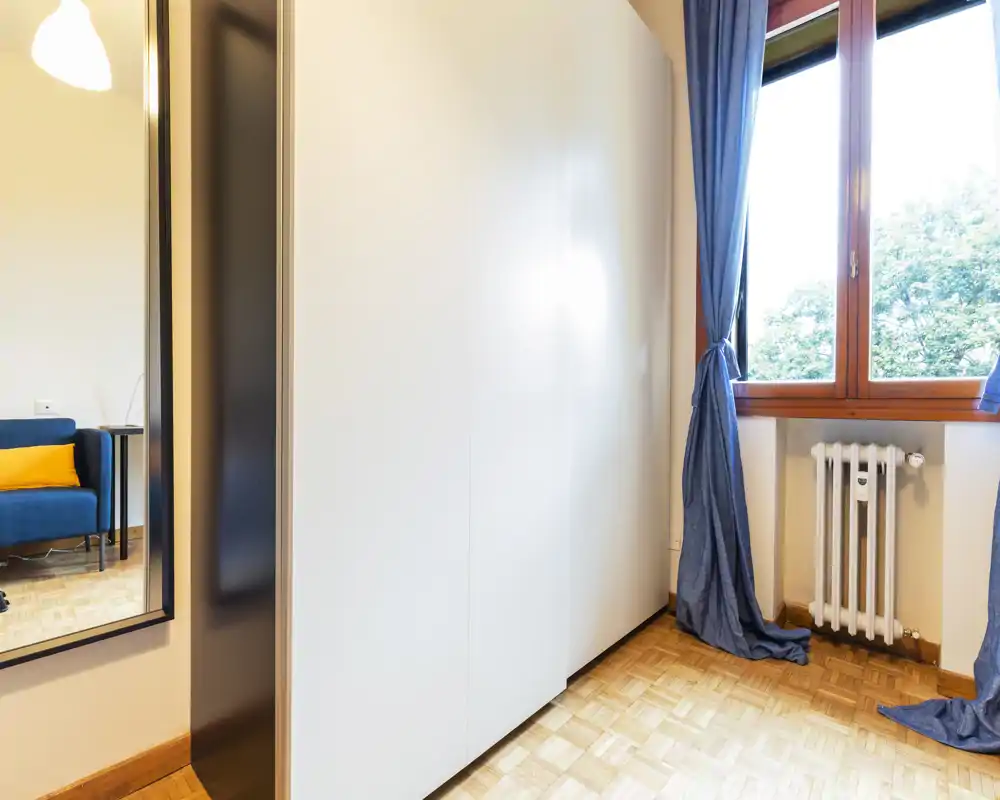 Coin salon lumineux avec une grande armoire et une fenêtre laissant entrer la lumière naturelle ; un canapé bleu avec un coussin orange est visible dans le miroir, suggérant un espace détente à proximité.