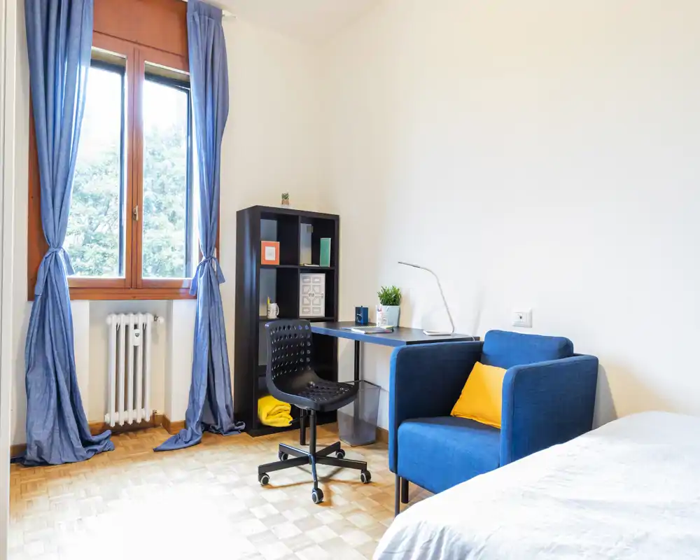 Coin chambre lumineux et moderne avec lit simple visible, fauteuil bleu et petit bureau près de la fenêtre. Luminosité naturelle, étagère sobre et touches chaleureuses.
