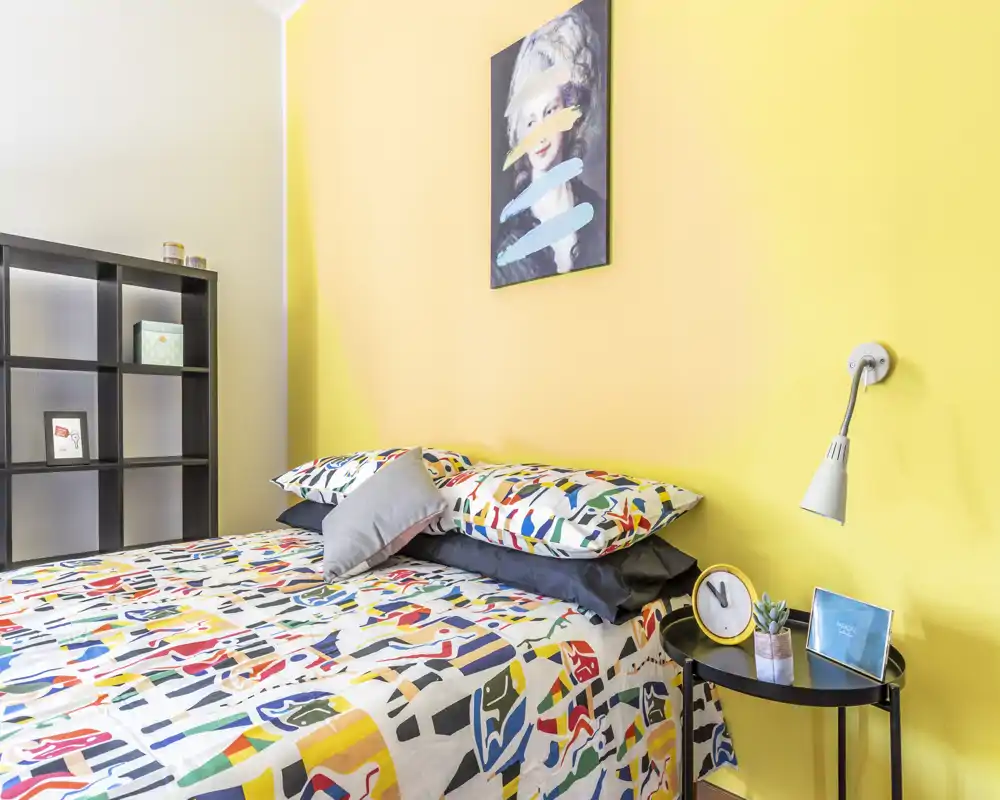 Bright, stylish bedroom with a colorful patterned bedspread, pillows and a bedside table with lamp and decor. The yellow feature wall and neat shelving add personality and a welcoming atmosphere.