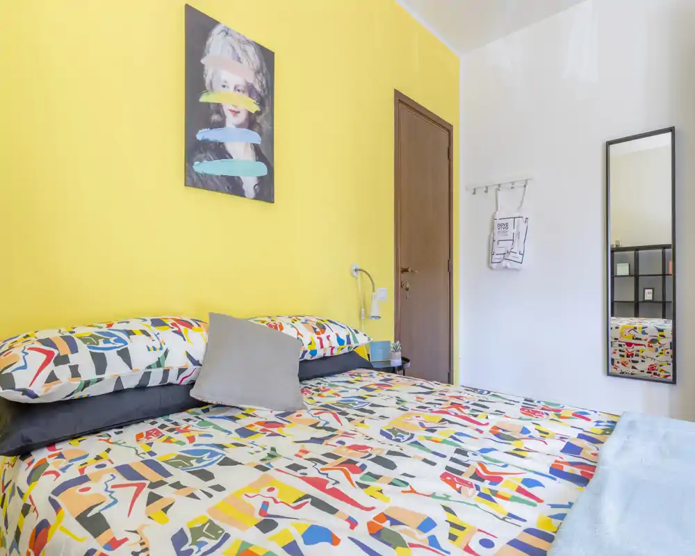 Bright, colorful bedroom with a patterned duvet and pillows against a cheerful yellow accent wall; bedside lamp and wall art create a cozy, well-styled space ideal for rental photos.