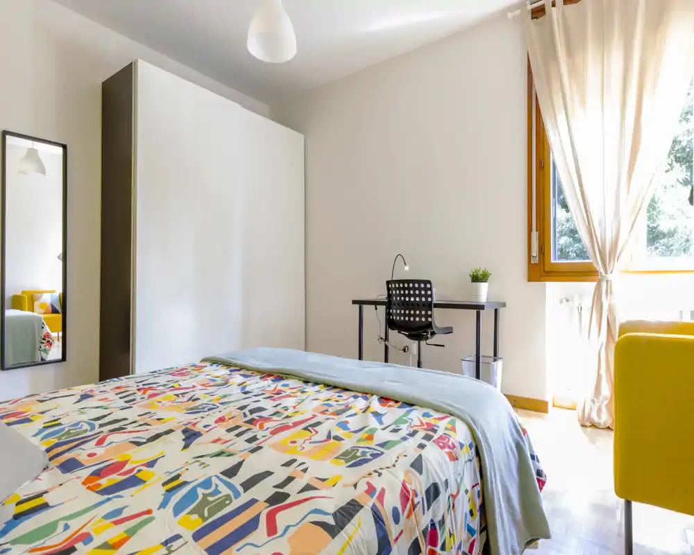 Bright, modern bedroom with a colorful patterned bedspread, a study desk by the window, wardrobe and natural light — cozy and well-lit for a restful stay.