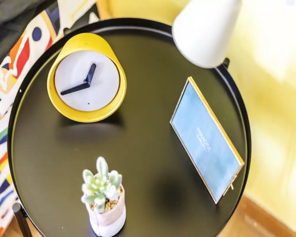 Close-up of a bedside table with a yellow clock, small succulent, framed card and lamp—stylish bedroom detail shot highlighting decor and lighting.