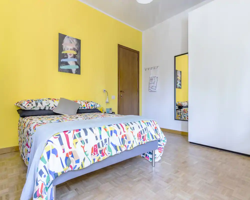 Bright, colorful bedroom with a double bed against a yellow accent wall, patterned bedding and simple modern furnishings — well lit and welcoming for guests.