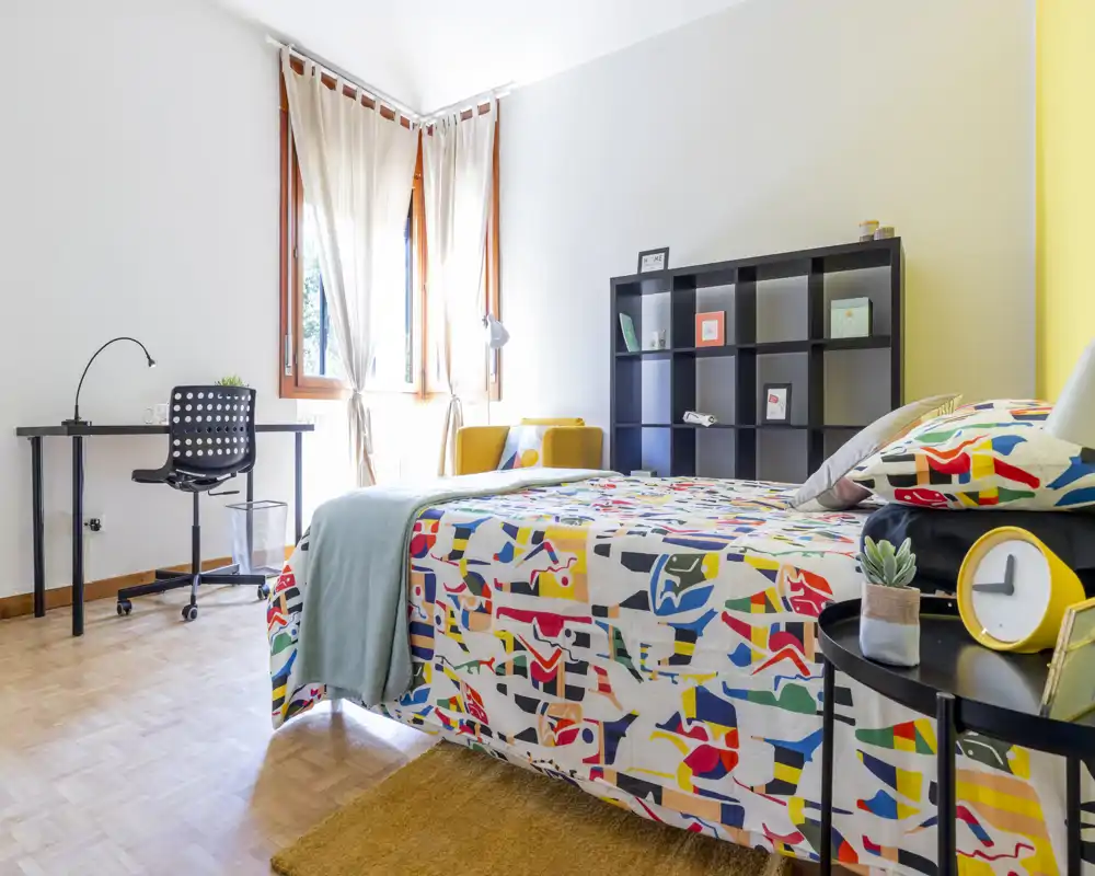 Bright, modern bedroom with a colorful patterned bedspread, bedside table with clock and plant, shelving unit and a study desk by the window. Natural light and warm wood flooring create a welcoming atmosphere.