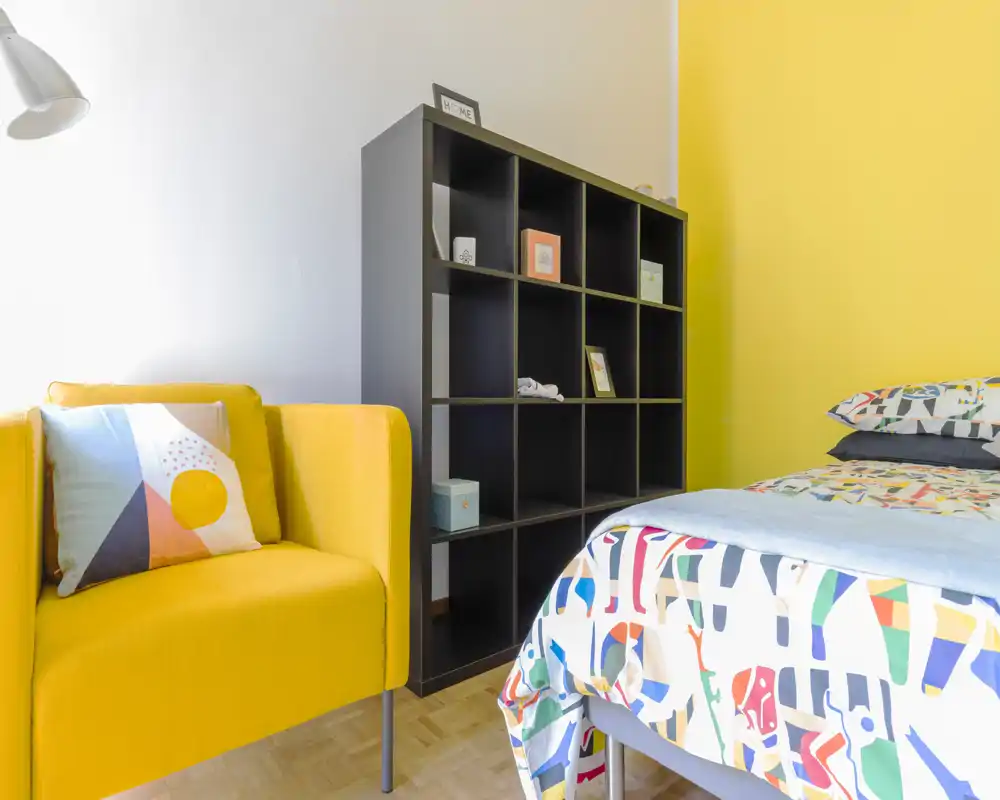 Bright, colorful bedroom corner with a patterned bed and a mustard-yellow armchair beside a black cubed shelving unit; modern, well-lit and thoughtfully styled for a welcoming feel.