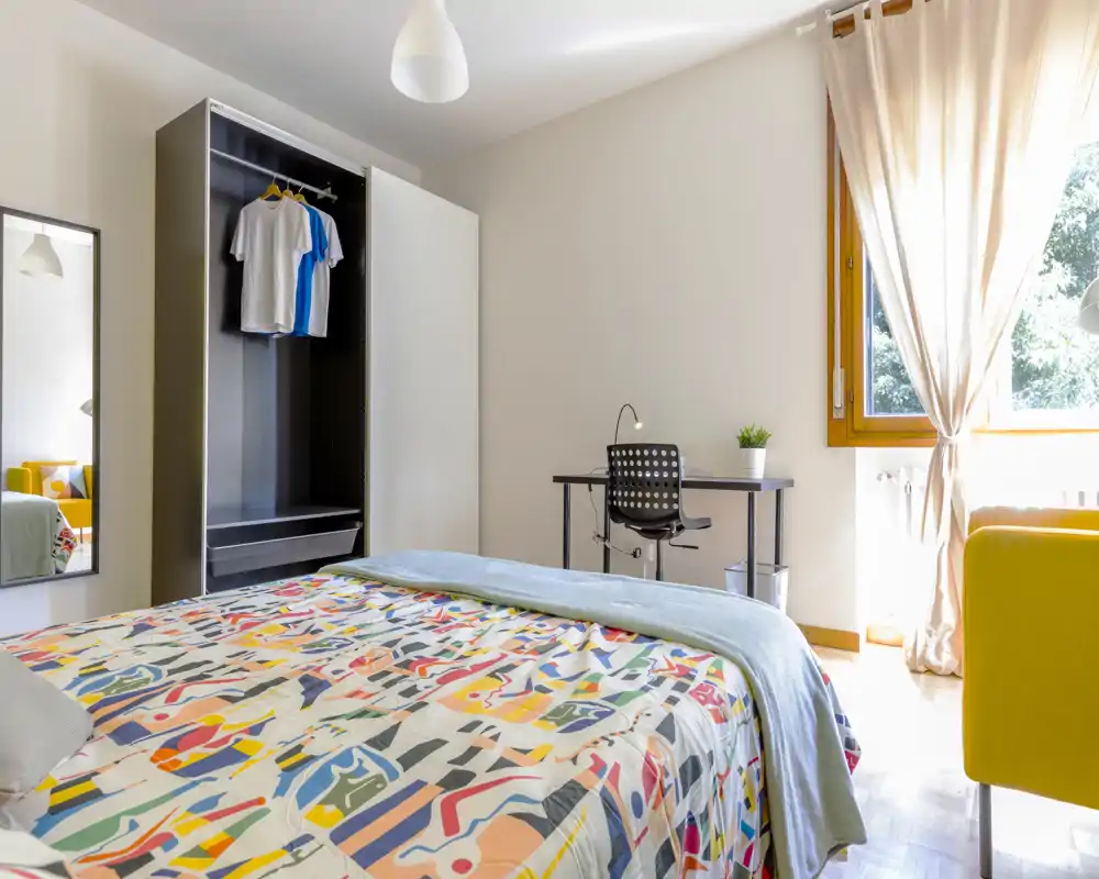 Bright, well-staged bedroom with a full bed, colorful bedding, a wardrobe, desk and a sunny window — ideal for rental listings highlighting natural light and a cosy atmosphere.