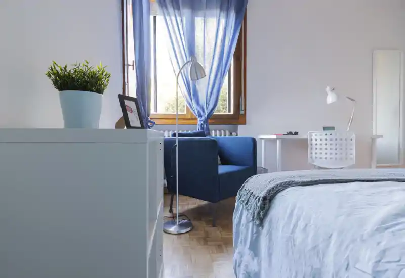 Bright, well-styled bedroom with a neatly made bed, a blue armchair by the window and a small desk — airy natural light and calm blue accents create a welcoming space.