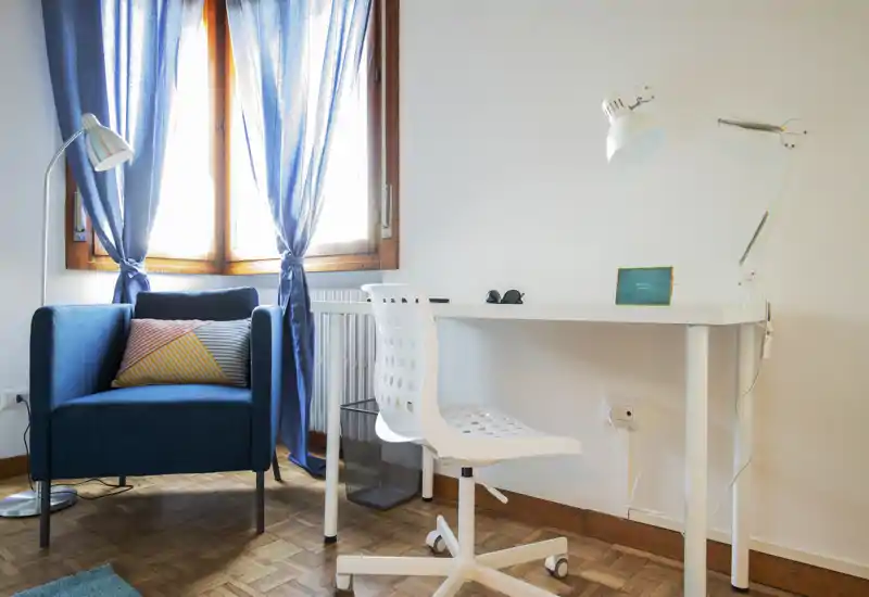 Bright, tidy living corner with a comfortable blue armchair, floor lamp and a white desk facing a sunlit window — ideal for relaxing or light work.