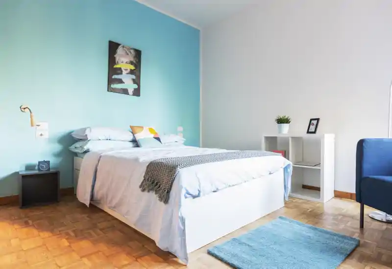 Bright, modern bedroom with a double bed against a turquoise accent wall, cozy textiles and simple shelving — ideal for relaxing stays.