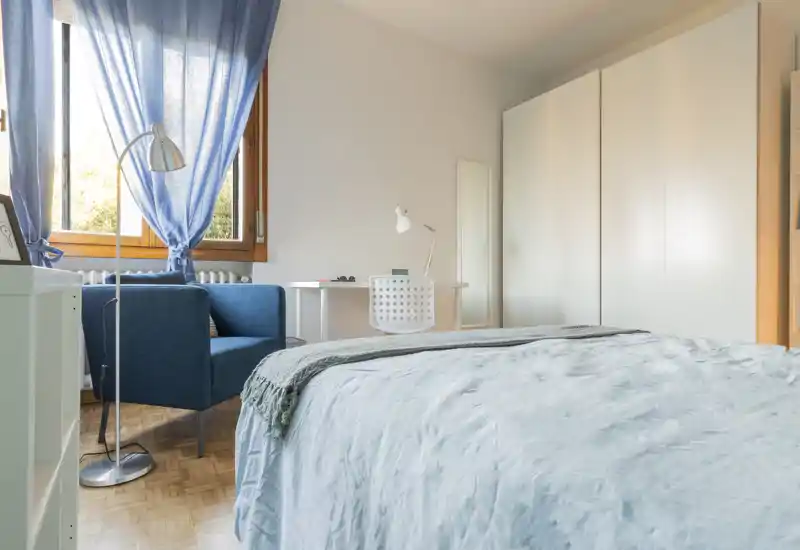 Bright, modern bedroom with a neatly made bed in the foreground, a blue armchair by the window, a small desk and large white wardrobe — ideal for rental listing photos.