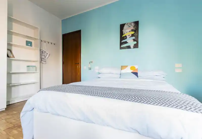 Bright, modern bedroom with a neatly made double bed against an aqua accent wall, decorative pillow and wall art, and built-in shelving offering storage.