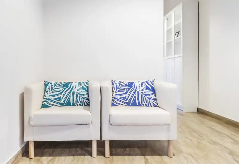 Minimal living area with two white armchairs and decorative blue and teal cushions, clean marble floor and bright neutral walls — ideal for a small lounge or waiting corner.