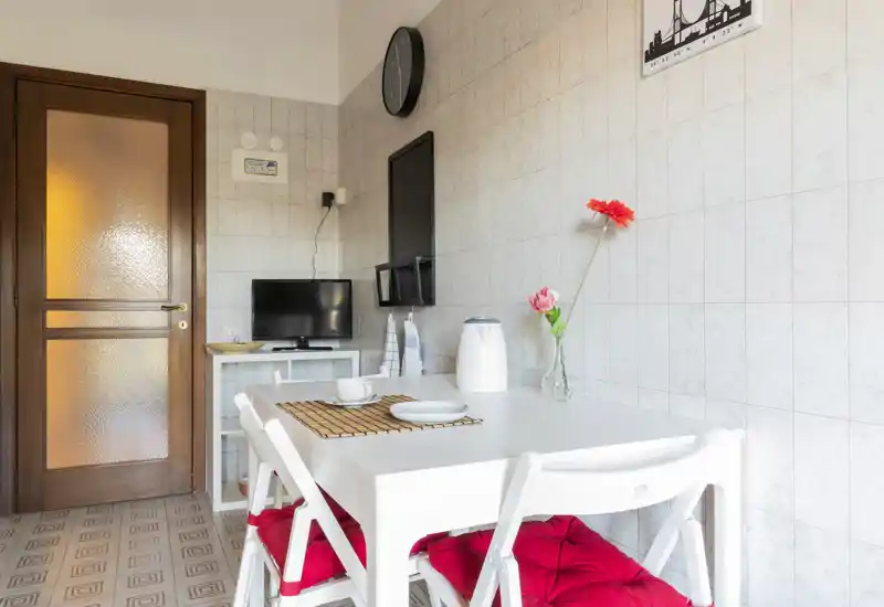 Compact, well-lit dining corner with a white table, folding chairs with red cushions, simple table setting and decorative flowers — ideal for a cozy two-person meal.