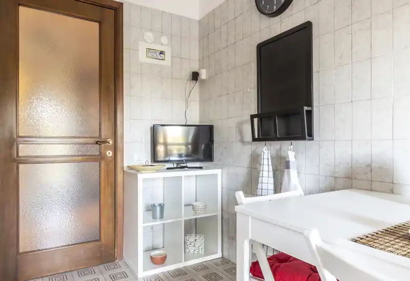 Compact living/dining corner with a white table, shelving unit and a small TV mounted on a tiled wall; light, tidy and simply decorated with towels and a red seat cushion.