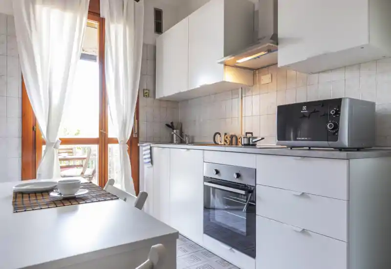 Bright modern kitchen with white cabinetry, built-in oven, microwave and a small dining table by a sunlit door leading to an outdoor area. Clean, airy space with light curtains and simple decorative touches.