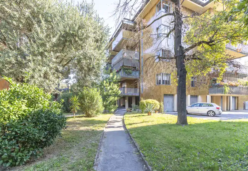 Well-maintained communal garden and entrance path in front of a residential building, with mature trees, shrubs and a small parking area — bright, peaceful outdoor space.