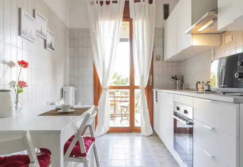 Bright, modern kitchen with white cabinets, built-in oven and a small dining table by a sunlit balcony door — cozy, well-lit and neatly styled.