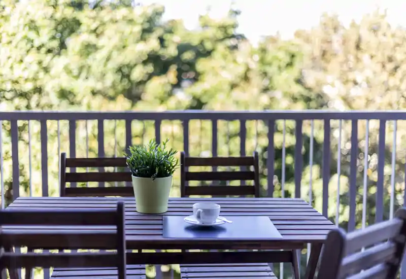 Sunny balcony with a wooden table and chairs set, a potted plant and a cup — ideal for morning coffee with leafy views.
