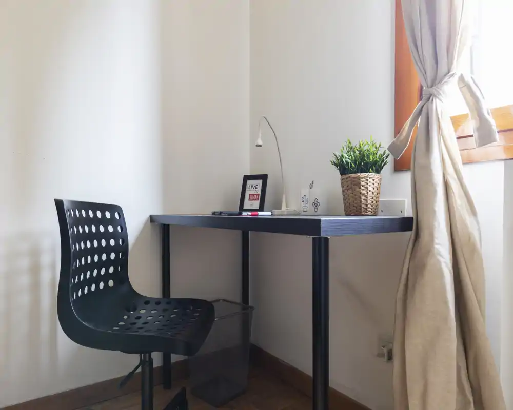 Bright, minimal study corner with a black desk and perforated chair by a window, styled with a potted plant and simple desk lamp.