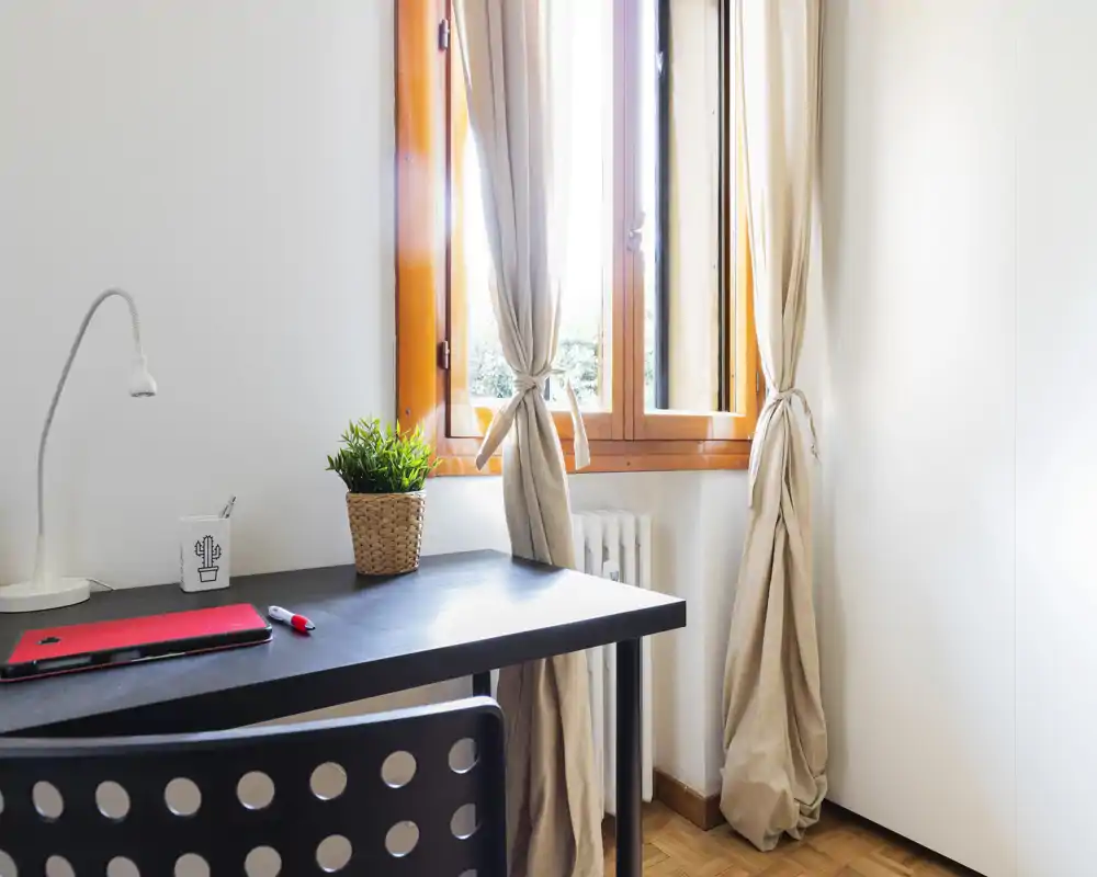 Bright corner workspace in a living area with a black desk, chair, task lamp and a window letting in natural light—simple, tidy and functional for remote work.