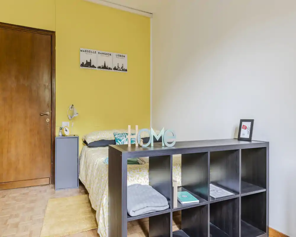 Sunny, well-staged bedroom with a double bed, cozy textiles and a low shelving unit separating the sleeping area; warm yellow accent wall and bright natural light make the space inviting.