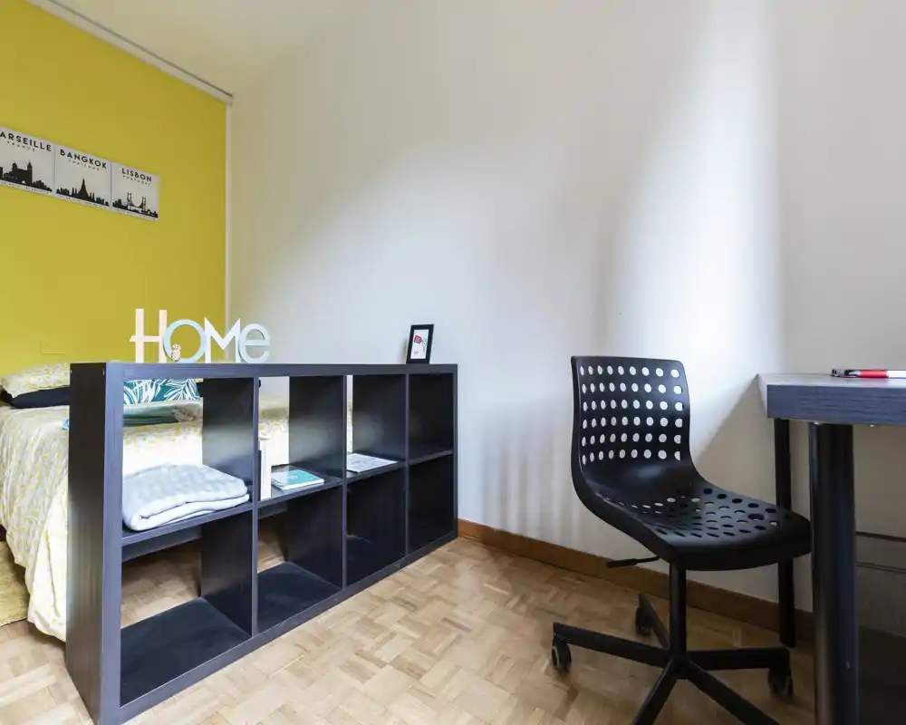 Bright, simply styled bedroom with a double bed partially visible behind a low shelving unit, wooden parquet floor, and a desk with a perforated chair — clean and welcoming.