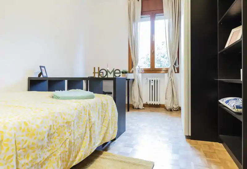 Sunny, tidy bedroom with a patterned yellow bedspread, open shelving as a headboard and a window providing natural light — a bright, welcoming sleeping space.