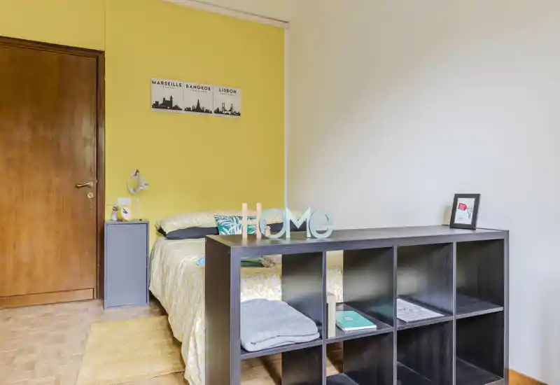 Sunny, well-staged bedroom with a double bed, cozy textiles and a low shelving unit separating the sleeping area; warm yellow accent wall and bright natural light make the space inviting.