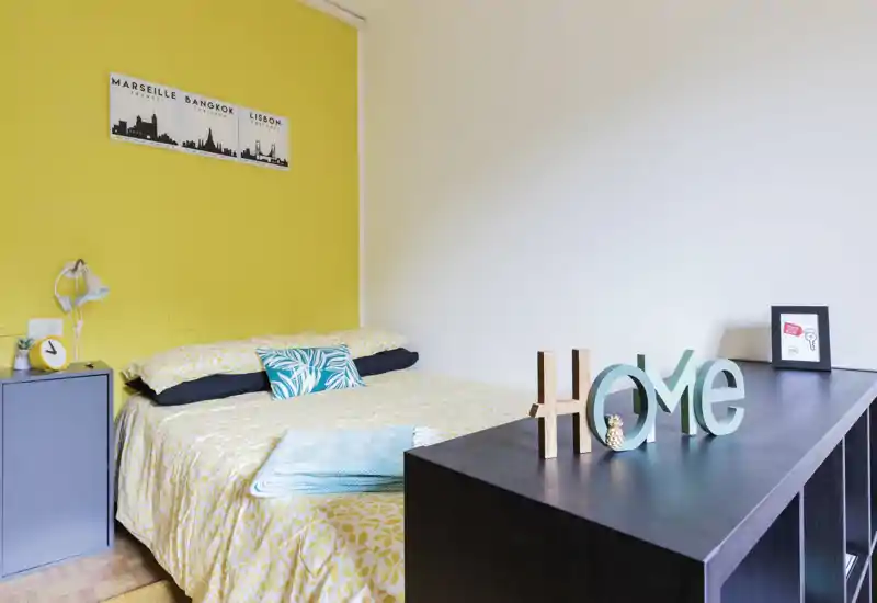 Sunny, well-styled bedroom with a double bed dressed in patterned yellow linens, decorative pillows and a bedside cabinet; a dark console with decorative 'HOME' letters frames the foreground.