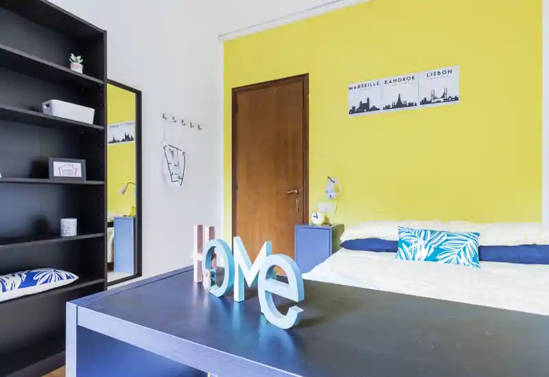 Bright, neatly staged bedroom with a double bed against a cheerful yellow accent wall, coordinated blue bedside table and decorative cushions creating a fresh, welcoming atmosphere.