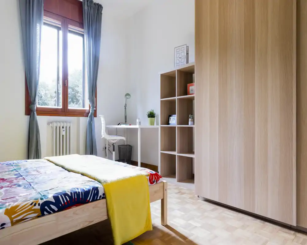Bright, tidy bedroom with a colorful bedspread, a wooden wardrobe and a small desk by the window — ideal for a single occupant or student.