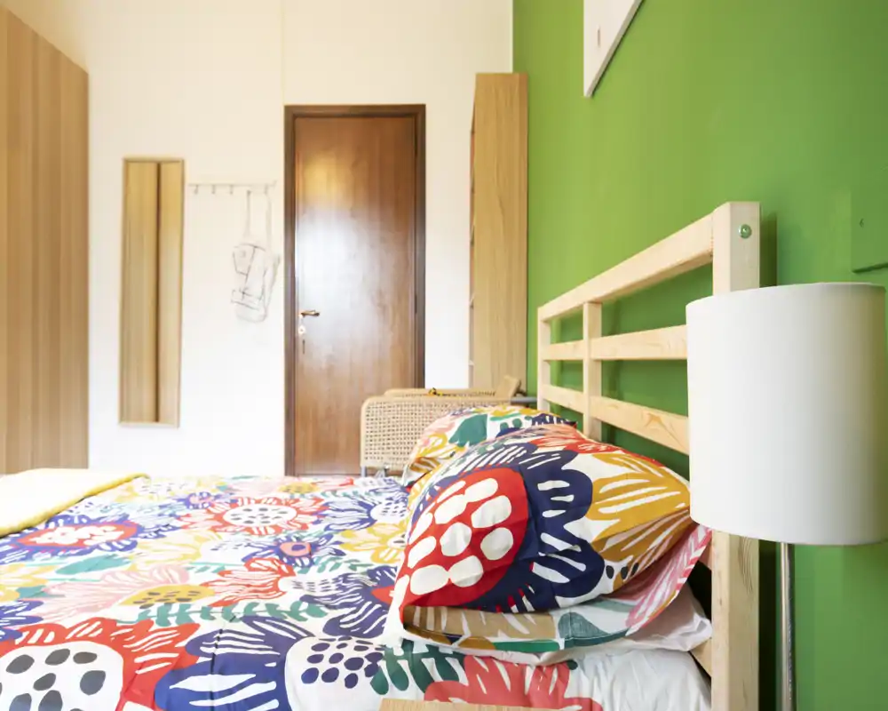 Bright, colorful bedroom with a wooden frame bed, patterned bedding and a bedside lamp. The image highlights a tidy double bed positioned against a green accent wall and warm wooden furniture.