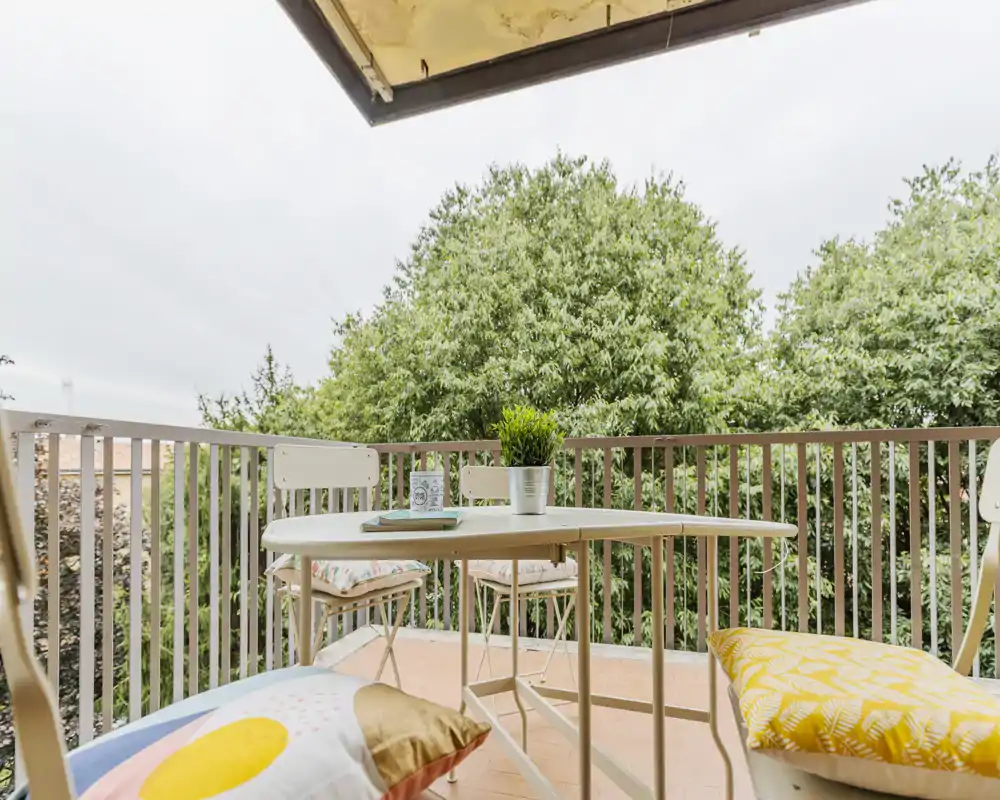 Sunny balcony with a small table, chairs and cushions overlooking leafy trees — a cozy spot for morning coffee or reading.