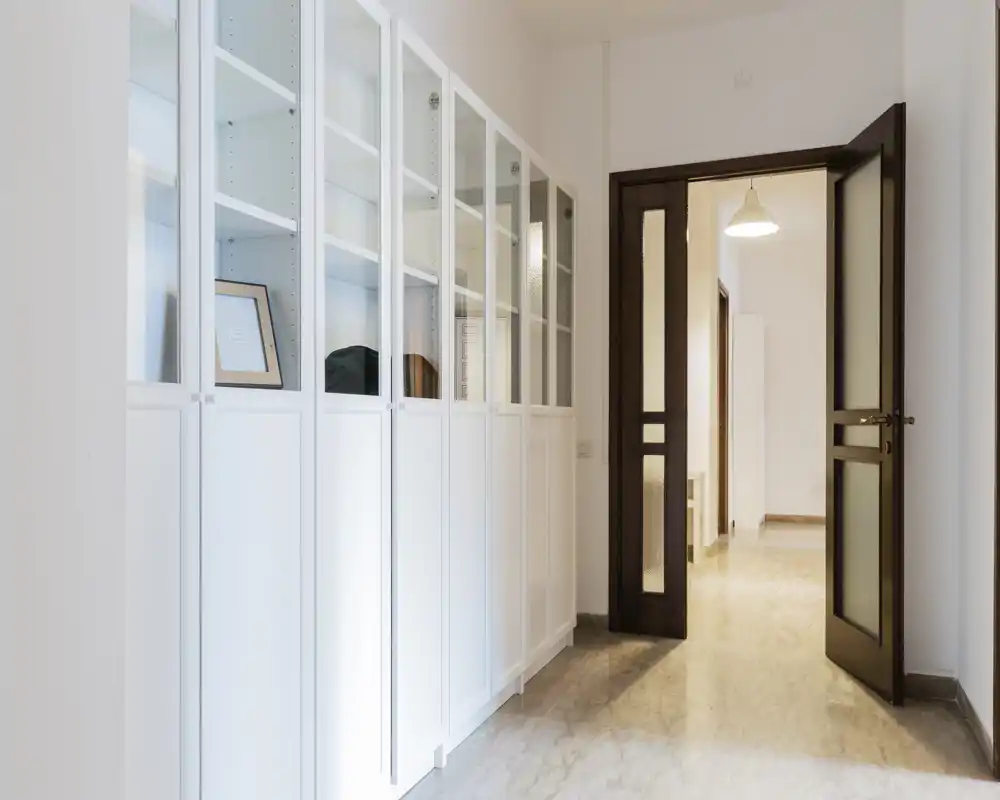 Bright, minimal entrance hallway with built-in white cabinets and a wooden door leading to the inner rooms. Marble-look floor and soft overhead lighting create a clean, welcoming first impression.