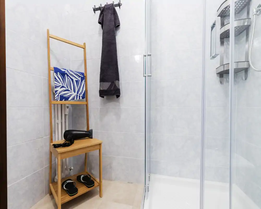 Modern, compact bathroom with a glass shower enclosure, towel hooks, and a wooden shelving unit holding a hairdryer and slippers.
