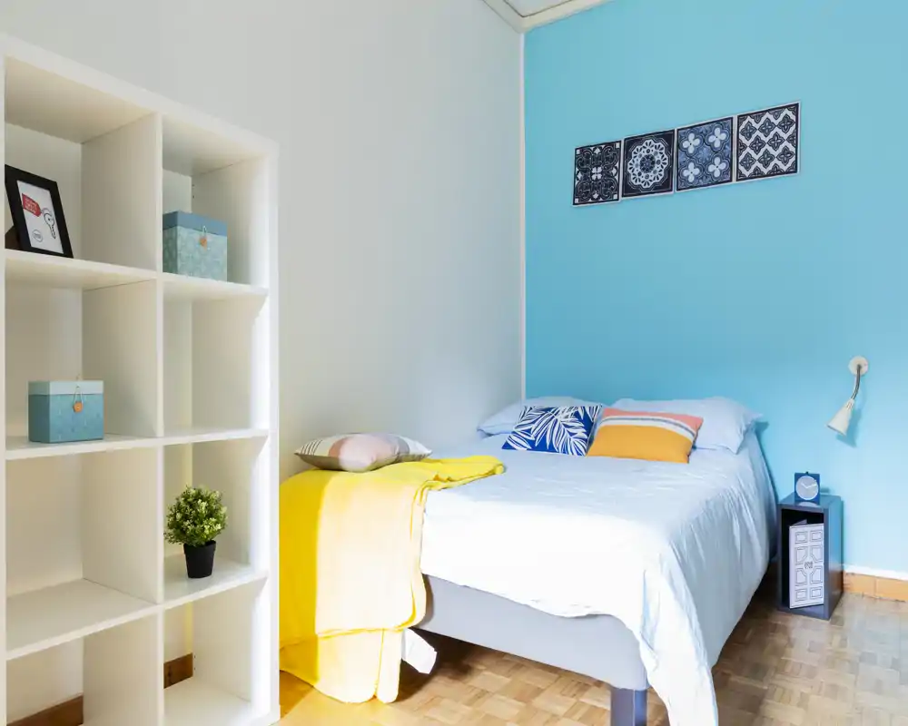Bright, neatly staged bedroom with a comfortable double bed against a turquoise accent wall, colorful pillows and a yellow throw, plus shelving for storage — ideal for rental photos.