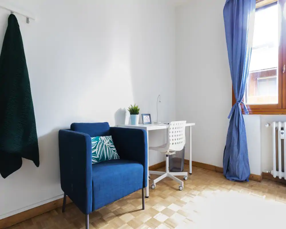 Bright, cozy living corner with a deep-blue armchair, patterned cushion and a small desk by a sunlit window.