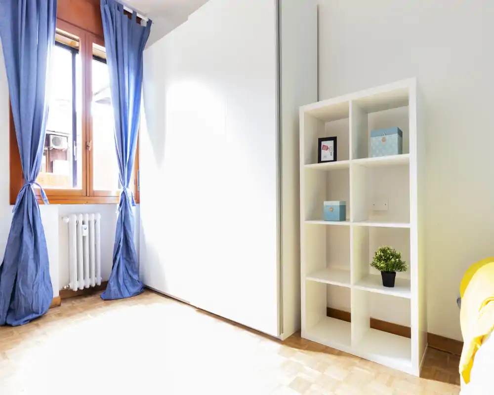 Bright, minimal bedroom corner with a white wardrobe, shelving unit and blue curtains. Sunlight fills the space creating a clean, airy feel—ideal for listing a single-bedroom or guest room.