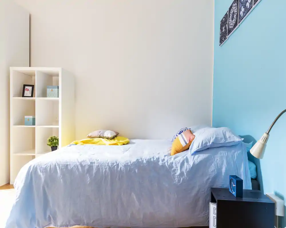 Bright, tidy single bedroom with a neatly made bed, colourful cushions and a small shelving unit. The room features a pleasant blue accent wall and natural light.