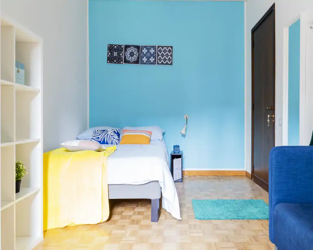Bright, styled bedroom with a double bed against a turquoise accent wall, colorful cushions and a yellow throw, wooden parquet floor and a small bedside lamp.