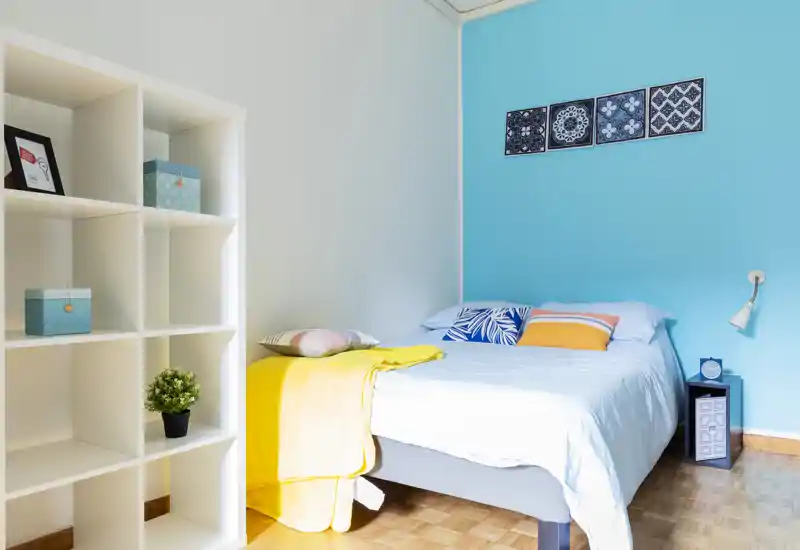 Bright, neatly staged bedroom with a comfortable double bed against a turquoise accent wall, colorful pillows and a yellow throw, plus shelving for storage — ideal for rental photos.