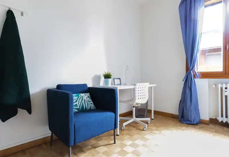 Bright, cozy living corner with a deep-blue armchair, patterned cushion and a small desk by a sunlit window.