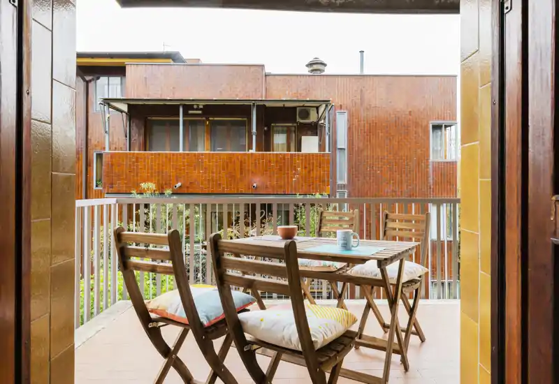 Sunny balcony with a small wooden dining set and cushions, overlooking neighboring brick buildings — ideal for morning coffee or an evening drink.