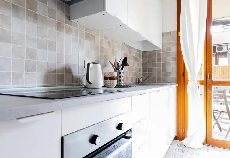Bright modern kitchenette with white cabinets, an induction cooktop, built-in oven and a sink by a large glazed door that opens to an outdoor seating area.