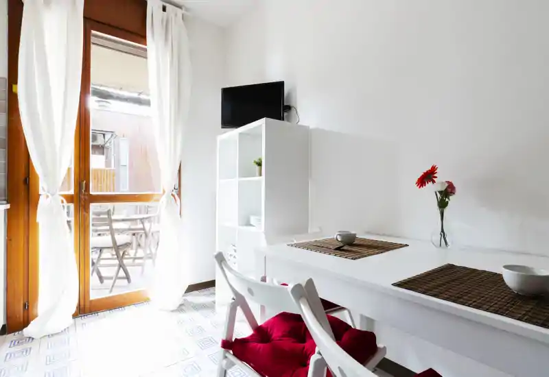 Bright, airy living/dining corner with a white table, red-cushioned chairs and a glass door opening to a small balcony. Minimal, fresh styling with natural light and simple decorative touches.