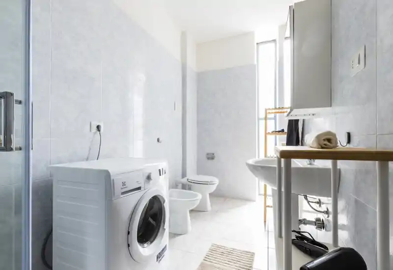 Bright, clean bathroom with a front-loading washing machine, sink, bidet and toilet; tiled walls and a tall narrow window provide natural light.