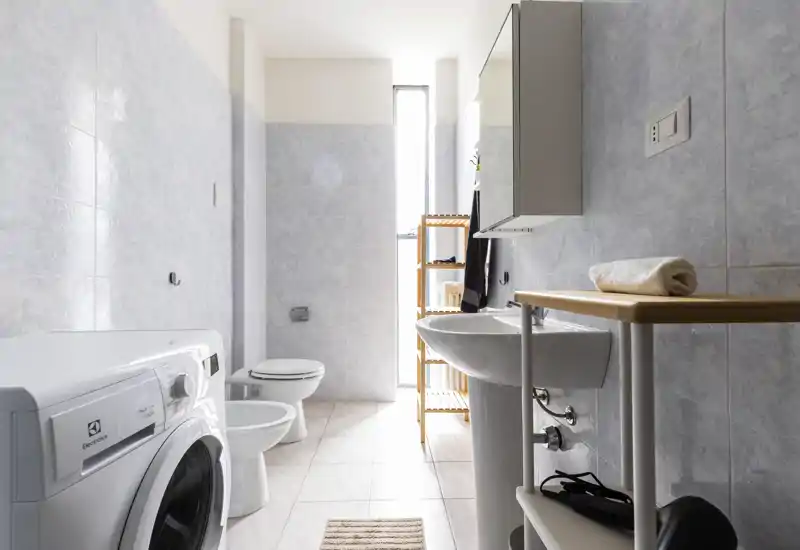 Bright, modern bathroom with white fixtures, a pedestal sink, toilet, bidet and a front-loading washing machine; light tiles and a tall narrow window provide natural light.