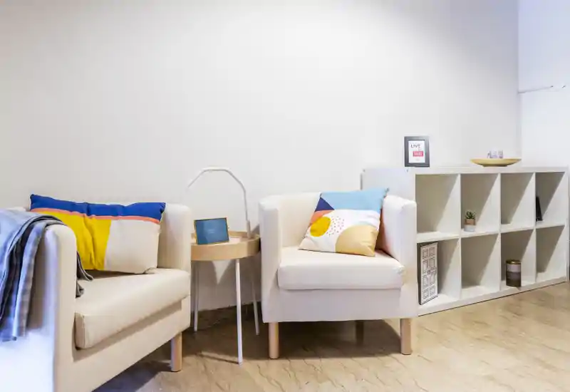 Bright, modern sitting area with two armchairs, colorful cushions and a small shelving unit — a cozy, well-lit living corner ideal for relaxing.