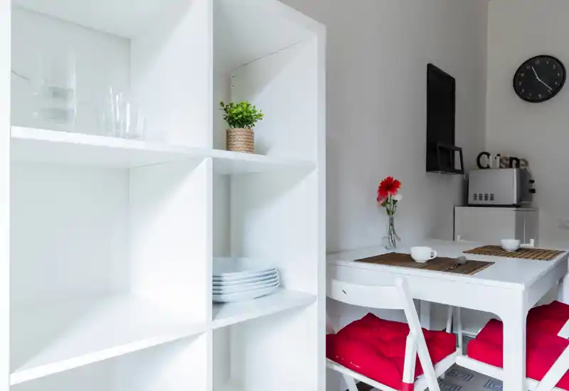 Bright, modern dining nook with a small white table set for two, red seat cushions, decorative placemats and a vase with a red flower; clean shelving and minimalist appliances visible in the background.